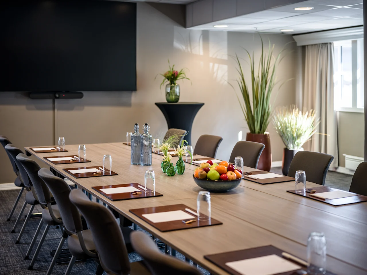 Ett modernt konferensrum med ett långt bord dukat för ett möte, med en stor skärm, frukt och växter.