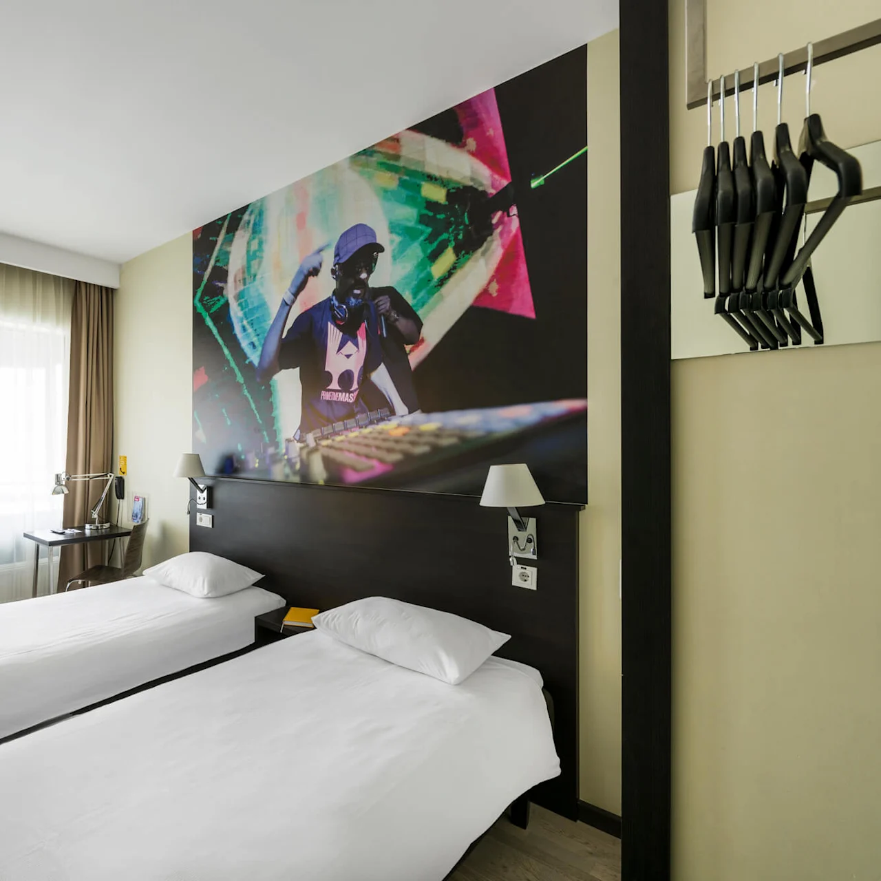Två prydliga enkelsängar vetter mot en väggmålning av en DJ i ett modernt hotellrum. En TV visar alternativen: "TRIP ADVISOR," "FACEBOOK," och "INSTAGRAM." En stol och ett skrivbord med lampa finns i närheten.