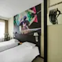 Two neatly made single beds face a wall mural of a DJ in a modern hotel room. A television displays options: "TRIP ADVISOR," "FACEBOOK," and "INSTAGRAM." A chair and desk with lamp are nearby.