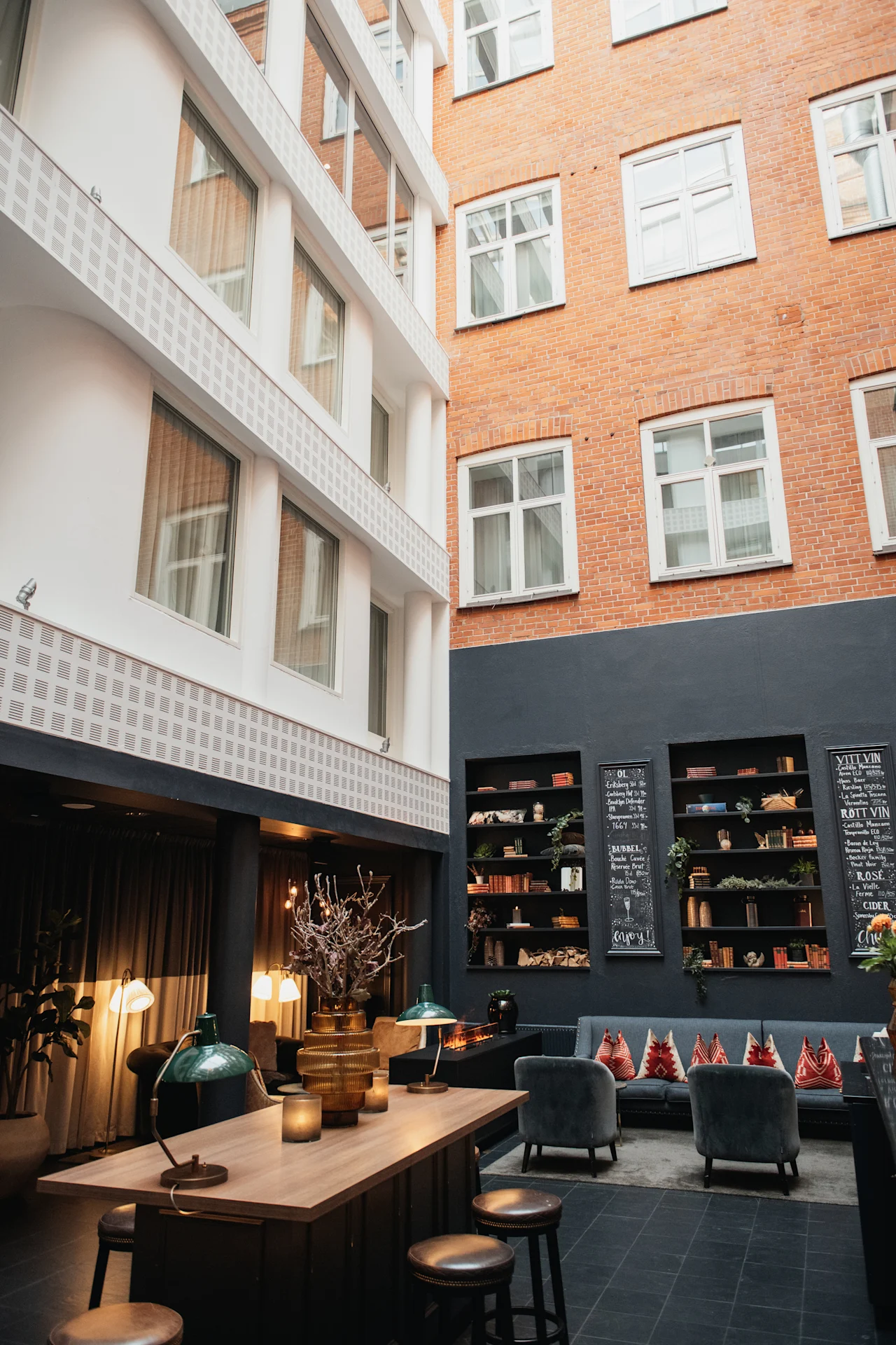 En indbydende hotellounge med et langt bord, komfortable siddepladser, bogreoler og tavler, der viser forskellige drikkevarer. Tekst: enjoy.