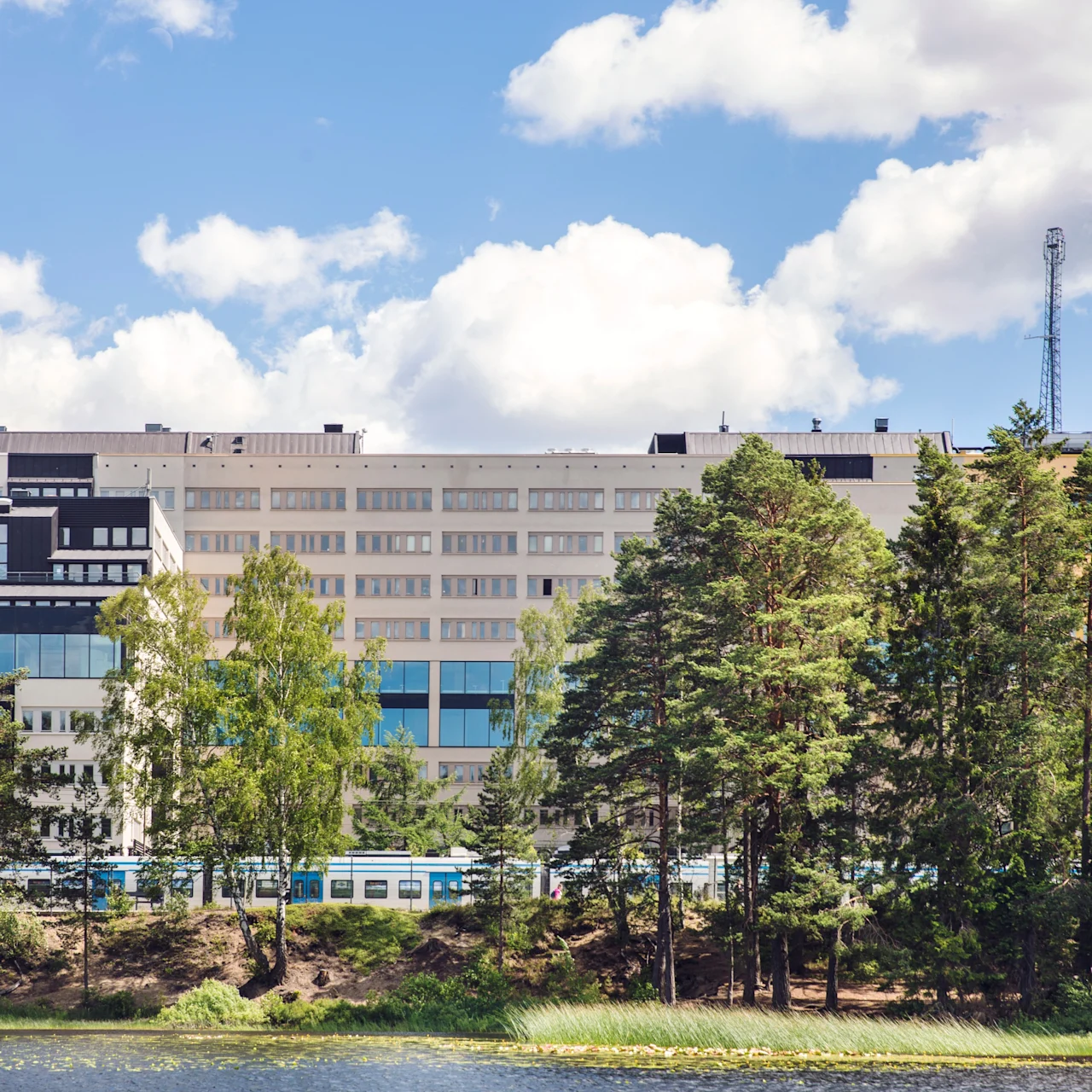 En stor, modern byggnad står framträdande bakom en rad höga träd, omgivet av en lugn vattensamling under en delvis molnig himmel.