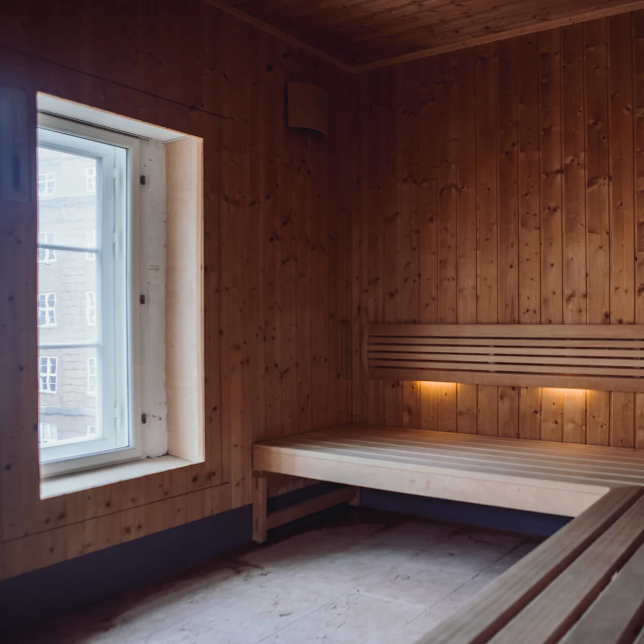 A wooden sauna room featuring benches with soft lighting underneath. A window on the left allows natural light, revealing an outside view of buildings.