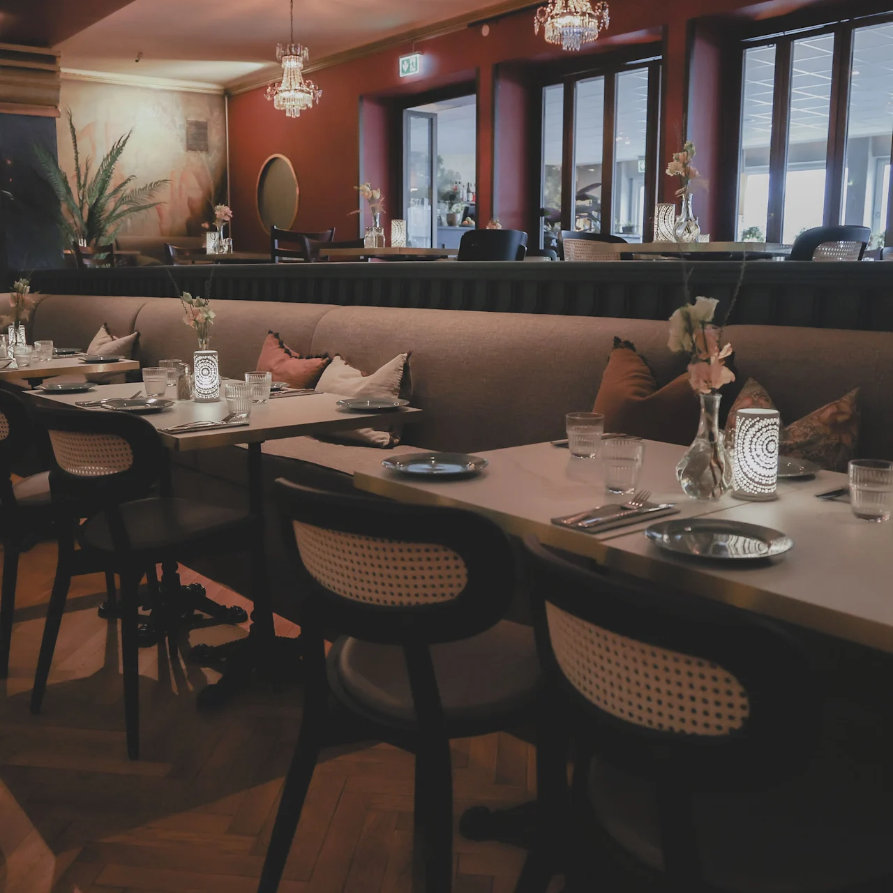 A long table is elegantly set with cutlery, glasses, and decorative flowers; chairs are neatly aligned. The restaurant interior features warm lighting and comfortable seating.