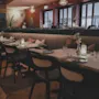 A long table is elegantly set with cutlery, glasses, and decorative flowers; chairs are neatly aligned. The restaurant interior features warm lighting and comfortable seating.