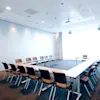 U-formede konferenceborde med tomme stole, placeret under et loft med indbygget belysning, i et lyst rum med hvide vægge og lukkede gardiner.