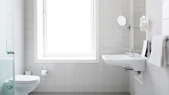 Puhtaan valkoinen kylpyhuone, jossa on vasemmalla wc-istuin ja pyyhe, oikealla pesuallas ja peili. Ikkuna tuo luonnonvaloa laatoitettuun tilaan.
