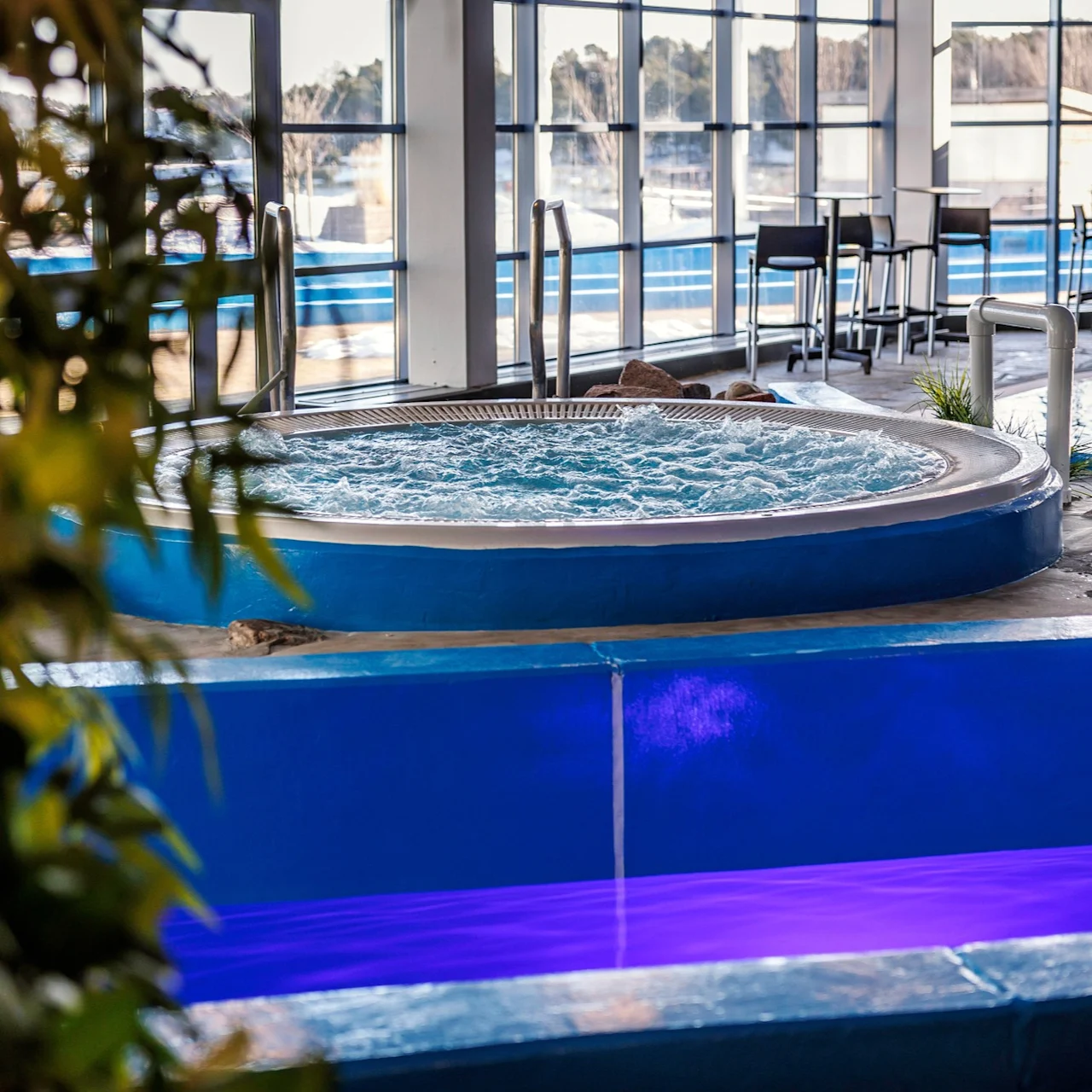 A circular hot tub with bubbling water is located indoors, surrounded by green plants and overlooking a windowed wall. Nearby are tables and chairs in a leisure area.