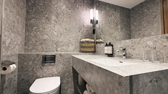 Ett modernt badrum med grå marmorliknande kakel, med toalett, handfat med tvål och glas, samt prydligt vikta handdukar.