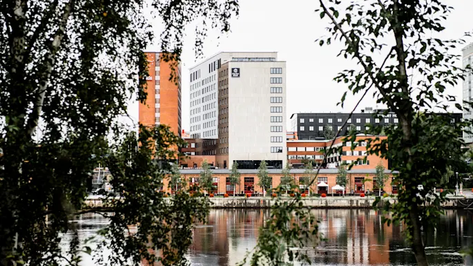 Høye bygninger står tvers over en elv, sett gjennom løvrike tregrener, og skaper et innrammet bylandskap.