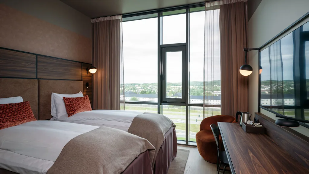 Et moderne hotellrom med to pent oppredde enkeltsenger, pyntet med røde puter, vender mot et stort vindu. Skrivebord i tre med stol og vannkoker ved siden av.