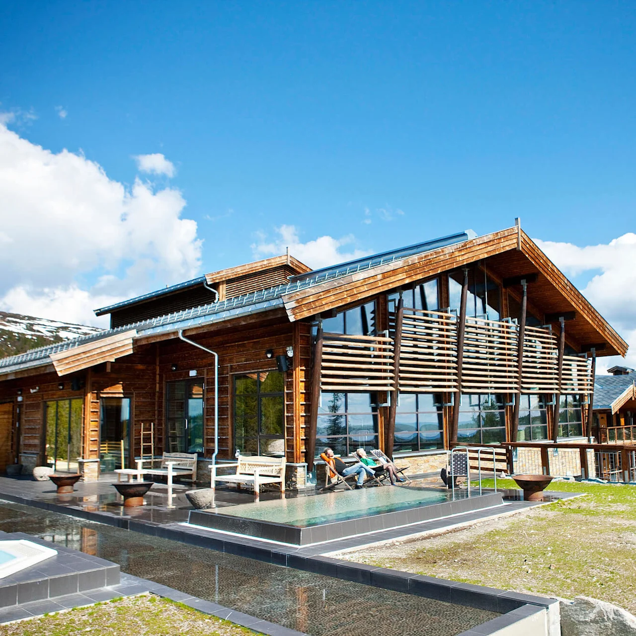 A modern wooden cabin with large glass windows rests in a mountainous landscape. People relax on deck chairs outside, enjoying sunny weather and clear blue skies.
