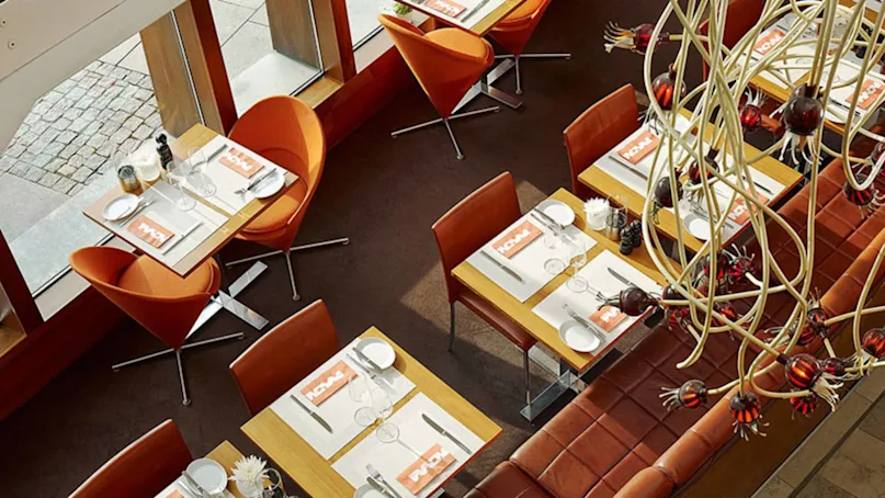 Restaurant tables are set for dining with white plates, glasses, and utensils on light wooden surfaces. Orange chairs and a decorative overhead chandelier create a stylish and inviting atmosphere. Napkins read "NOMA."