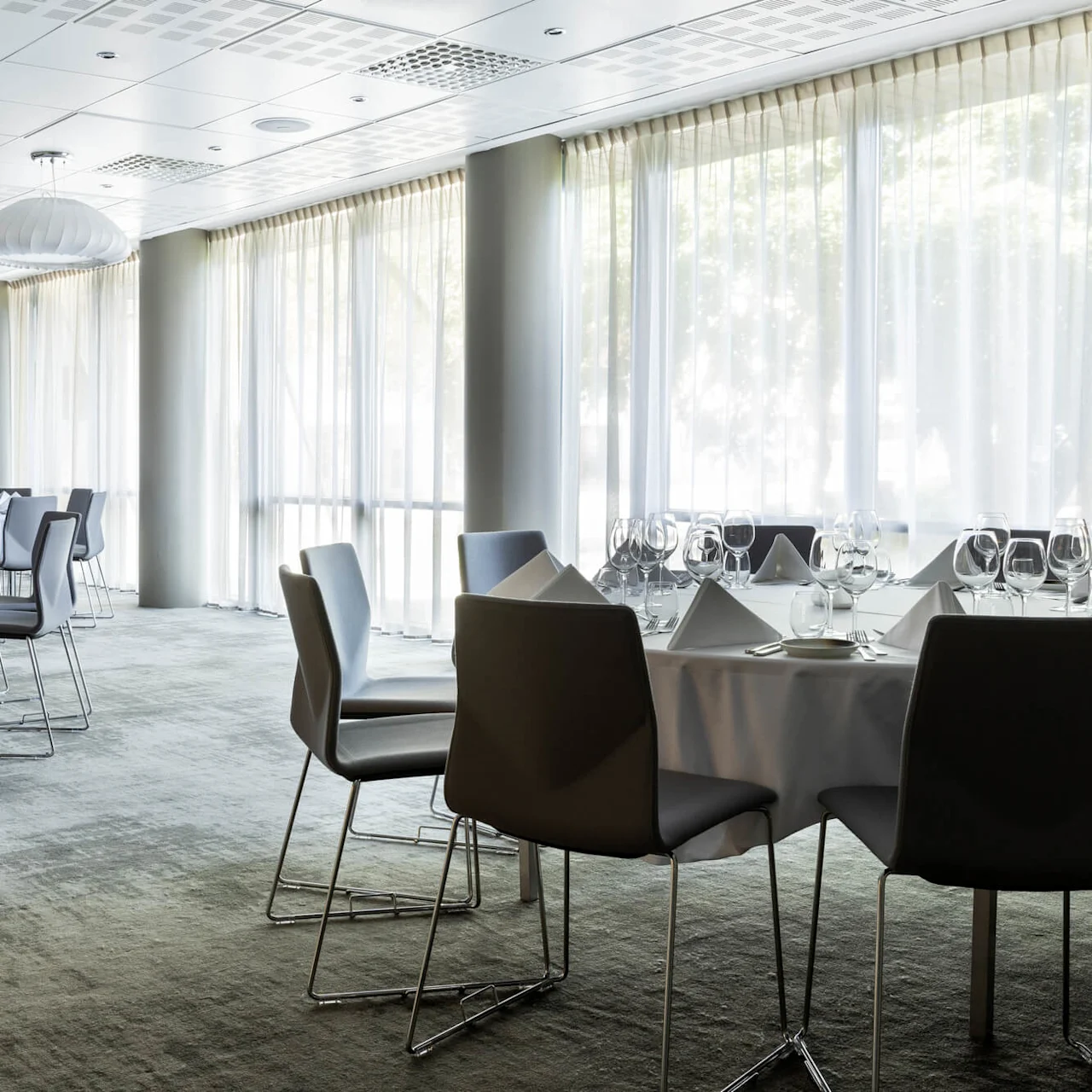 Tables are elegantly set with glassware and napkins, surrounded by chairs in a spacious room with large windows and soft natural light. No text present.