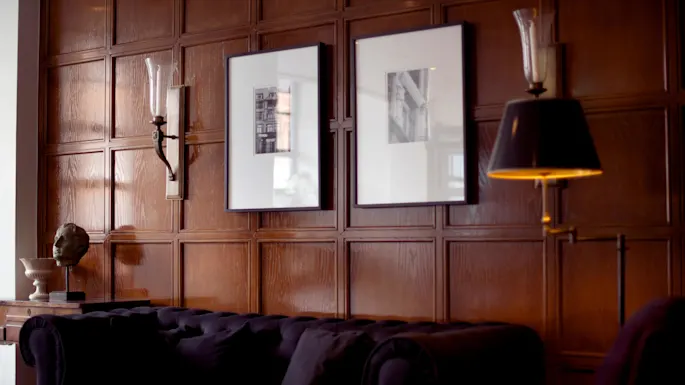 A dark-toned, elegant room with wall sconces, framed photographs, a wooden panel wall, and a tufted sofa partially visible.