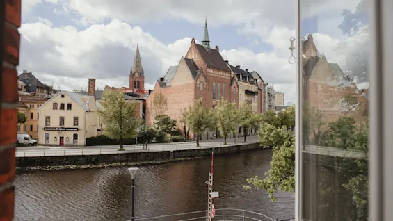 En tegelkyrka med spetsigt torn, reflekterad i ett fönster, står vid en trädbevuxen flodbank under molnig himmel. Närliggande byggnader förstärker stadens historiska landskap.