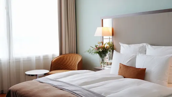 A neatly made bed is adorned with white pillows and a brown cushion, flanked by a bedside table with a flower vase, under soft lighting. Nearby, a chair and a window with curtains.