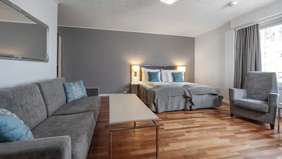 Et pænt indrettet hotelværelse har en dobbeltseng mod en grå væg, flankeret af lamper, med en grå sofa og lænestol på trægulv, og et vindue med gardiner.