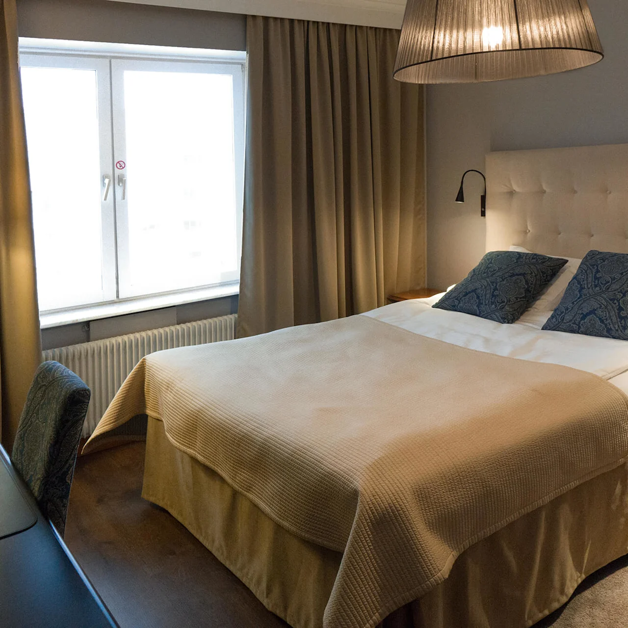 En seng med to blåmønstrede puter hviler under en stor lampe, omgitt av beige gardiner, et vindu og et skrivebord med en stol i et koselig rom.