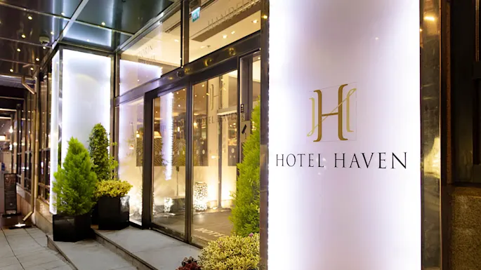 En hotellinngang har en glassdør omgitt av potteplanter. Skiltet sier "HOTEL HAVEN" i elegant skrift. Miljøet er opplyst og innbydende.