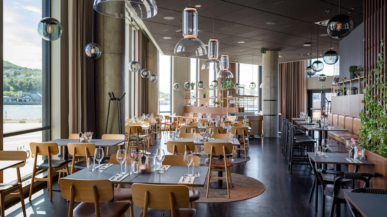 Et moderne restaurantinteriør med dækkede borde, store vinduer med udsigt over en strandpromenade og stilfulde pendellamper.