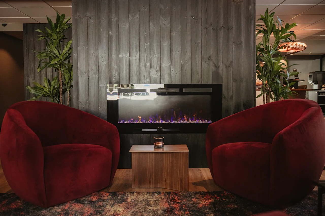 Two plush red armchairs invite relaxation in a cozy lounge with an electric fireplace and lush green plants.