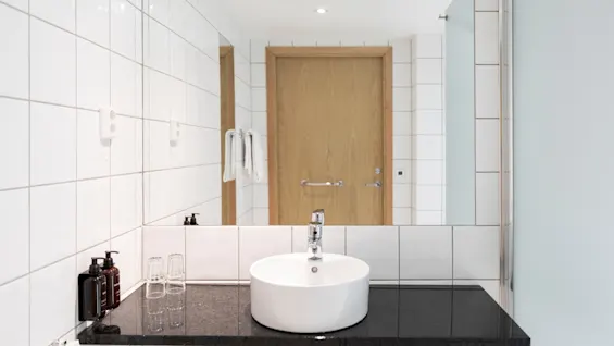 A clean and modern bathroom with a white sink, dark countertop, and a mirror reflecting a wooden door and fresh towels.