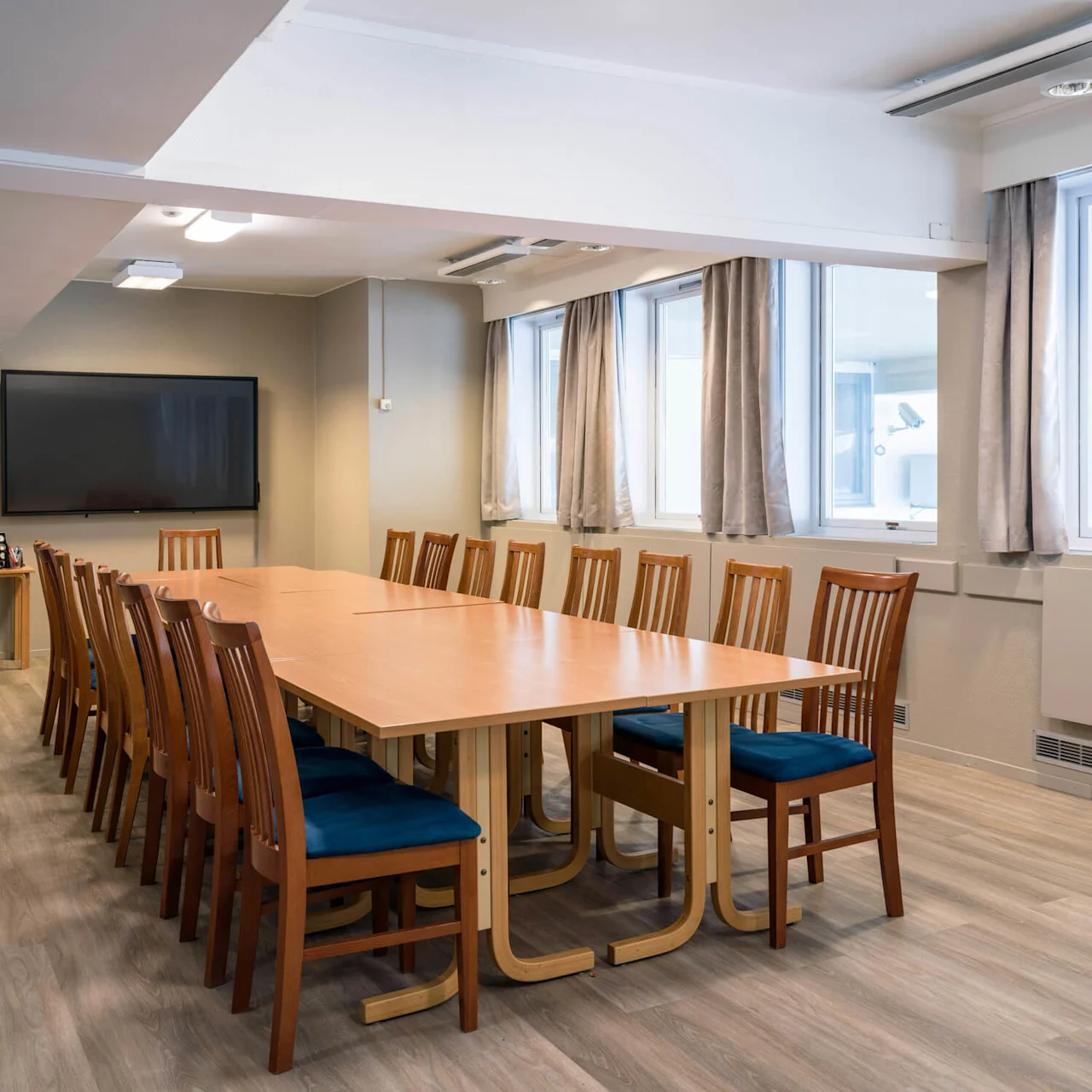 A long wooden conference table surrounded by chairs stands in a bright room with large windows and a wall-mounted television. Light wooden flooring and neutral curtains enhance the setting.