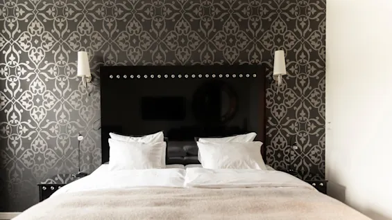 Tyylikäs hotellihuone, jossa on sänky valkoisilla vuodevaatteilla, tumma sängynpääty ja koristeellinen kuviotapetti.