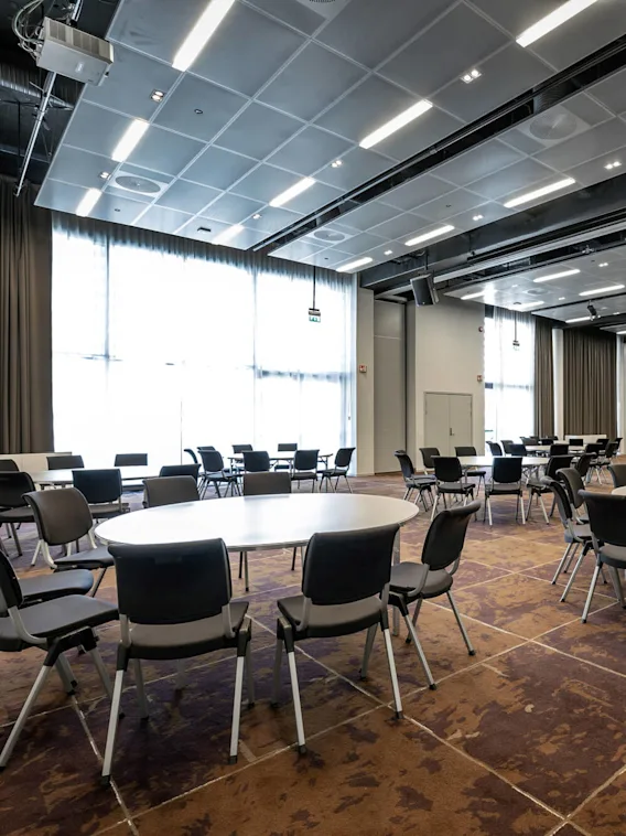 Runde bord med stoler arrangert i et romslig konferanserom; lyst opp av store vinduer, med et mønstret brunt gulv og moderne takarmaturer.