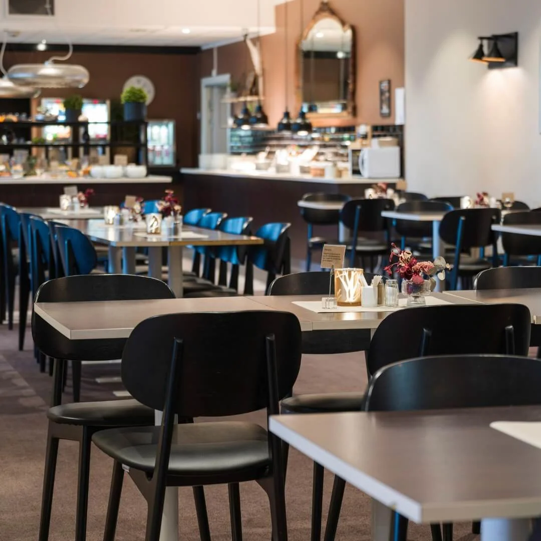 Borde placeret i en restaurant, pyntet med vaser og lys, omgivet af stole. En buffet er synlig i baggrunden med lys og dekorationer, der forbedrer den elegante spiseatmosfære.