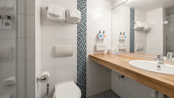Badeværelse med toilet, vask og træbænk. To sæbedispensere og håndklæder er pænt arrangeret. Hvide vægge med blå mosaik detaljer. Blødt lys.