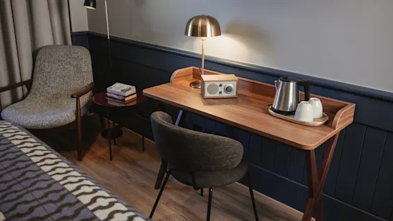 Ett mysigt hotellrum med ett träskrivbord med vattenkokare, koppar och en radio, tillsammans med en bekväm fåtölj och säng.