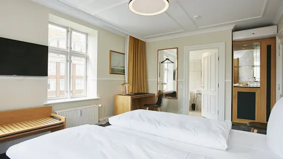 Et lyst og indbydende hotelværelse med en komfortabel seng, et skrivebord, et TV og et glimt af badeværelset.