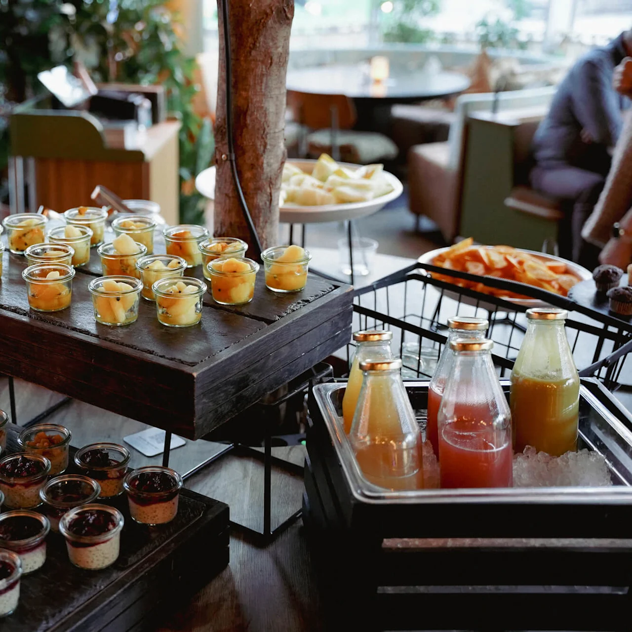En buffetopsætning med forskellige desserter i glaskopper og flasker juice på is, indendørs med gæster og grønt i baggrunden.