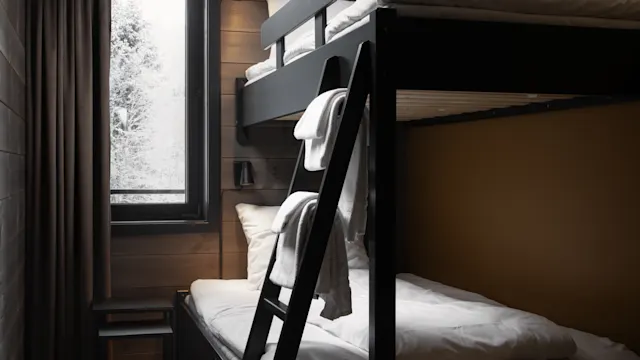 Bunk bed with white bedding stands against a brown wall. A window with curtains filters soft light, while a small stool sits nearby, creating a cozy, minimalist bedroom.