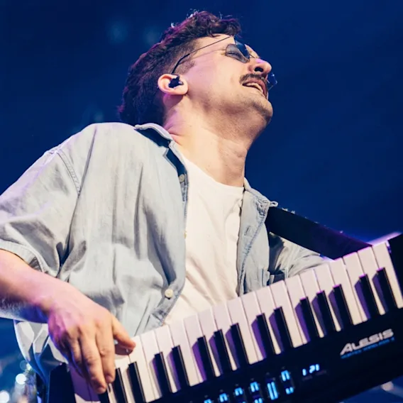 En mannlig musiker med solbriller som spiller på en svart keytar på scenen under blått konsertlys.
