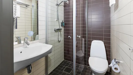 A small bathroom features a sink, shower with glass door, and toilet. Tiled walls are beige and brown. A towel rack holds folded towels above the sink.