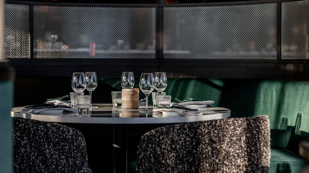 Ett runt matbord dukat med vinglas, servetter och bestick; omgivet av mjuka stolar i en svagt upplyst, mysig restaurangbås.