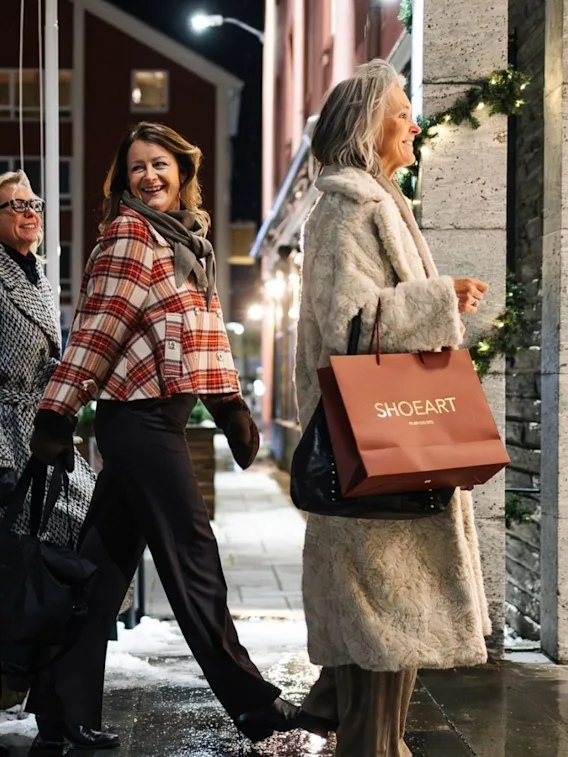 Tre personer går på en våt bygata om natten, en bærer handleposer, med festlige lys som pynter bygninger.