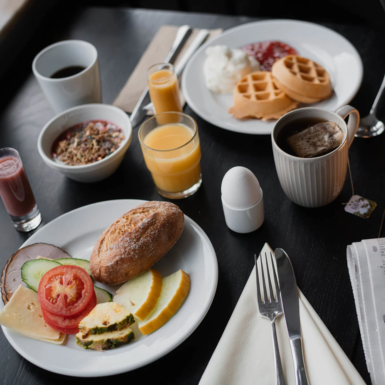 Et frokostbord med forskjellige retter inkludert vafler, frukt og egg. Drikkevarer inkluderer juice, kaffe og en smoothie. En avis ligger i nærheten, sammen med bestikk på et mørkt bord.