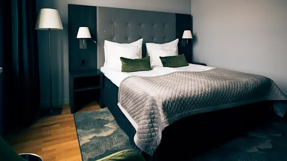 A neatly made bed with green cushions and a gray quilt, flanked by nightstands and wall-mounted lamps, is situated in a softly lit room with wooden floors.