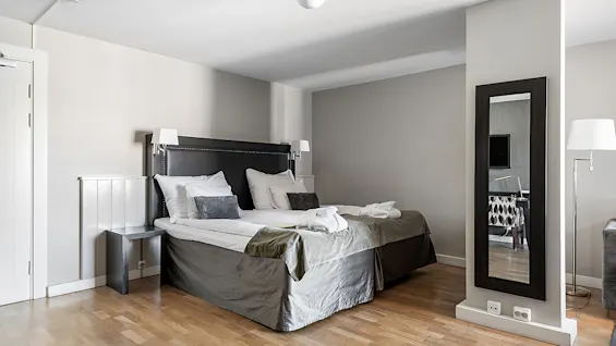 A neatly made bed with gray and white bedding in a clean, modern bedroom with wooden floors, white walls, and minimalistic furniture.