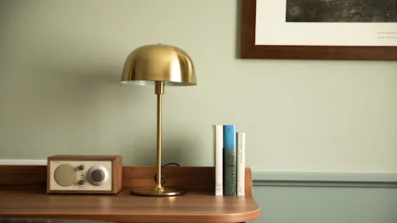 En mässingbordslampa står på ett träbord bredvid en vintage radio och tre böcker. En inramad bild hänger på den blekgröna väggen.