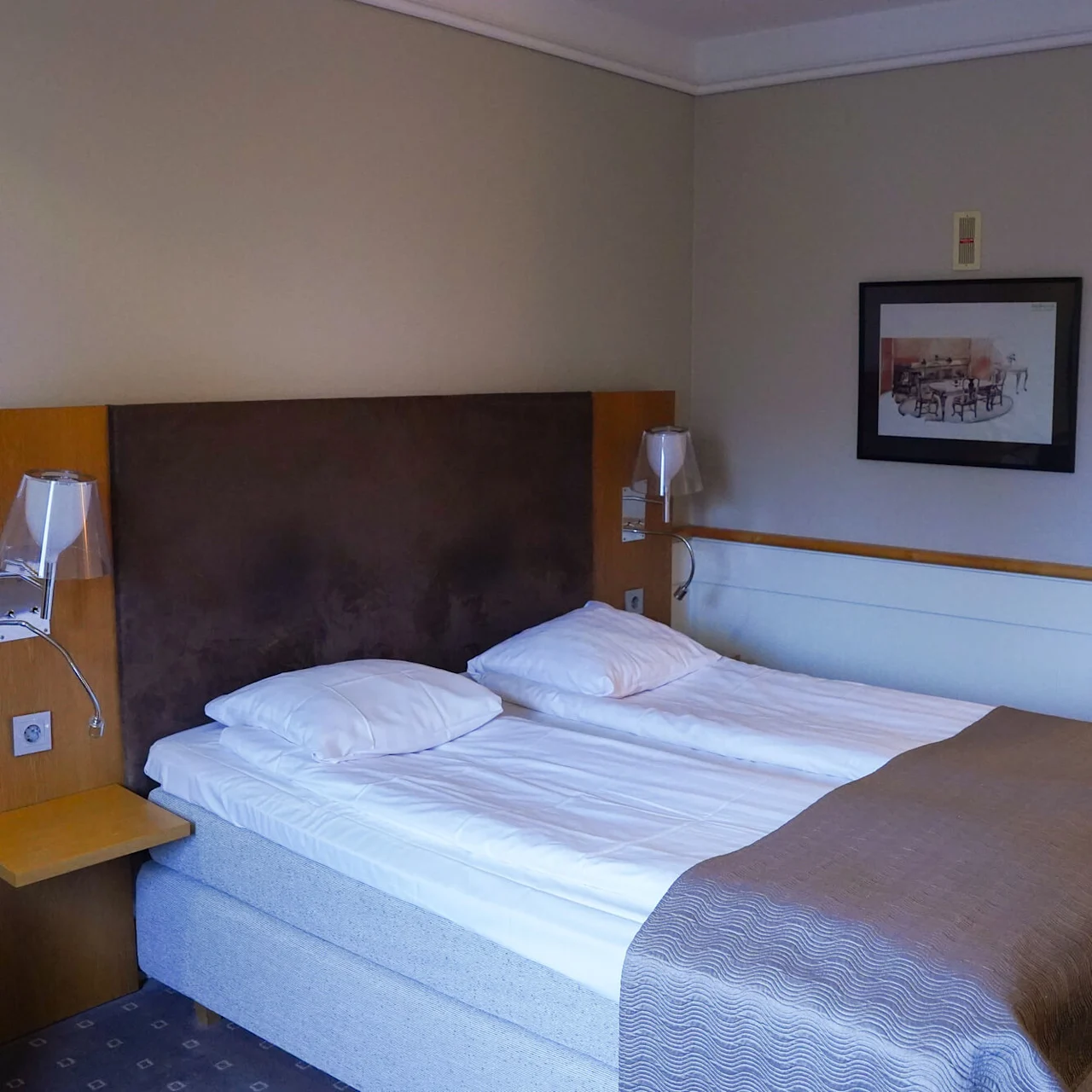 A neatly made twin bed in a hotel room with a wooden headboard, a nightstand, a wall lamp, and artwork.