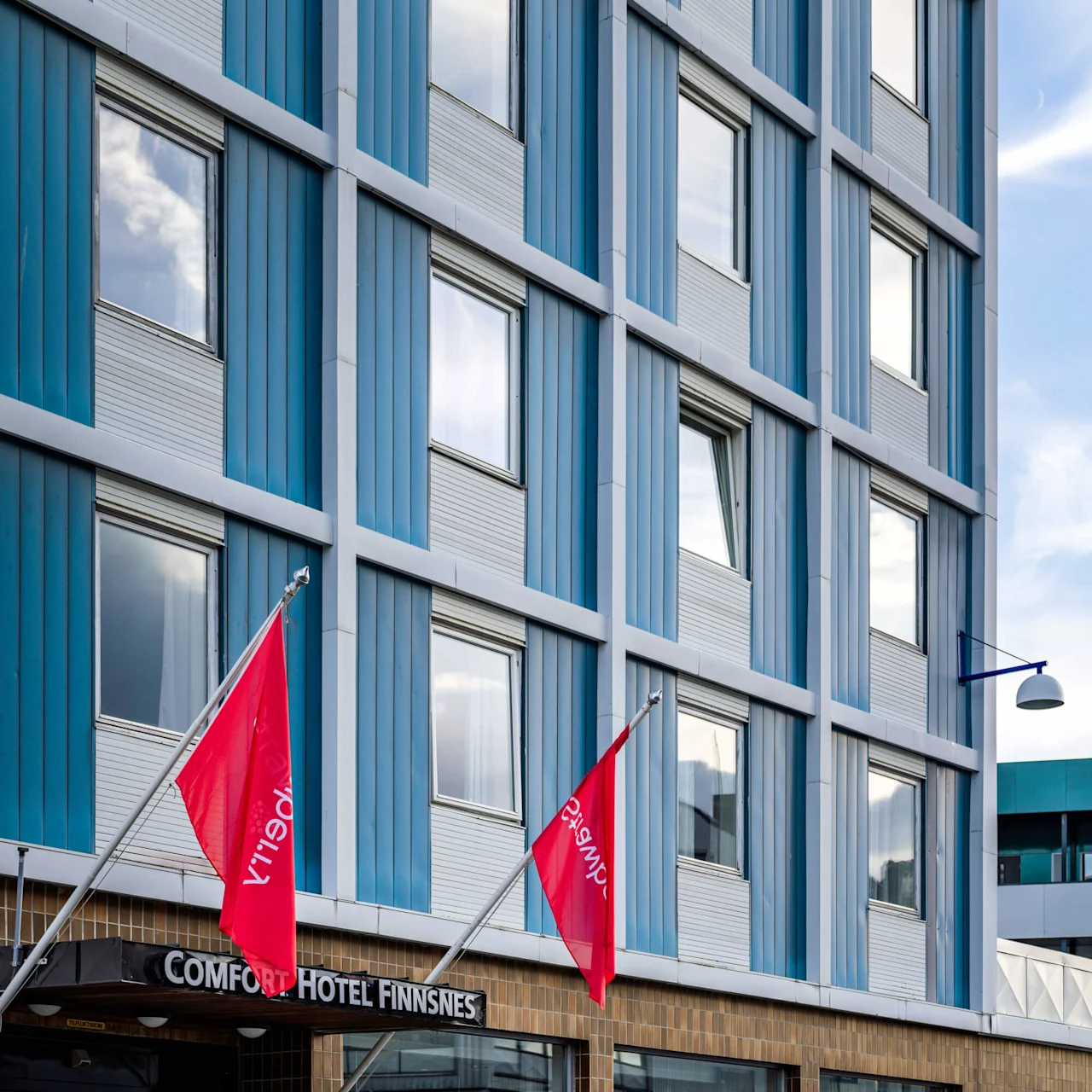 A hotel entrance with "COMFORT HOTEL FINNSNES" sign, flanked by two red flags and white chairs. Surrounding glass windows reflect the cloudy sky.