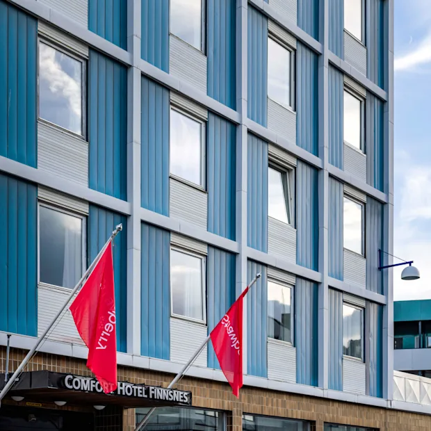 En hotelindgang med skiltet "COMFORT HOTEL FINNSNES", flankeret af to røde flag og hvide stole. Omgivende glasvinduer reflekterer den overskyede himmel.