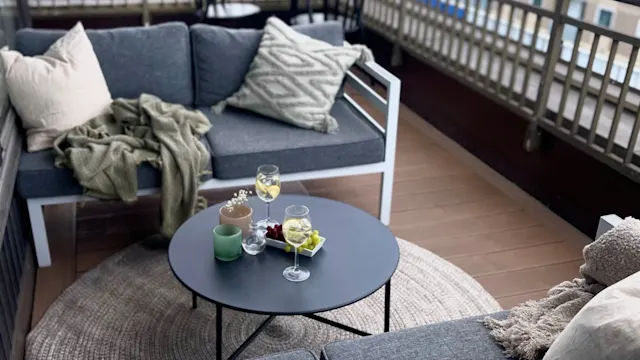 A gray sectional sofa on a balcony holds cushions and a blanket. A round table displays drinks and snacks. The balcony overlooks trees and buildings.