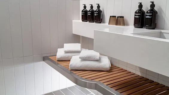 White towels rest on a wooden bench in a modern bathroom. Five bottles labeled "GROWN ALCHEMIST" and two cups are arranged on a white countertop.