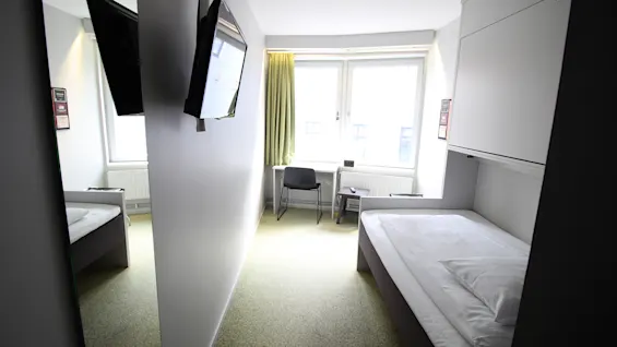 Valoisa ja kompakti hotellihuone, jossa on yhden hengen sänky, pieni työpöytä ikkunan ääressä ja seinälle asennettu tv.