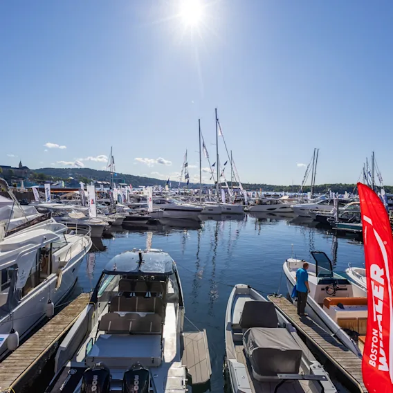 En solrik dag i en travel marina med mange båter fortøyd, og en person som står på et av fartøyene.
